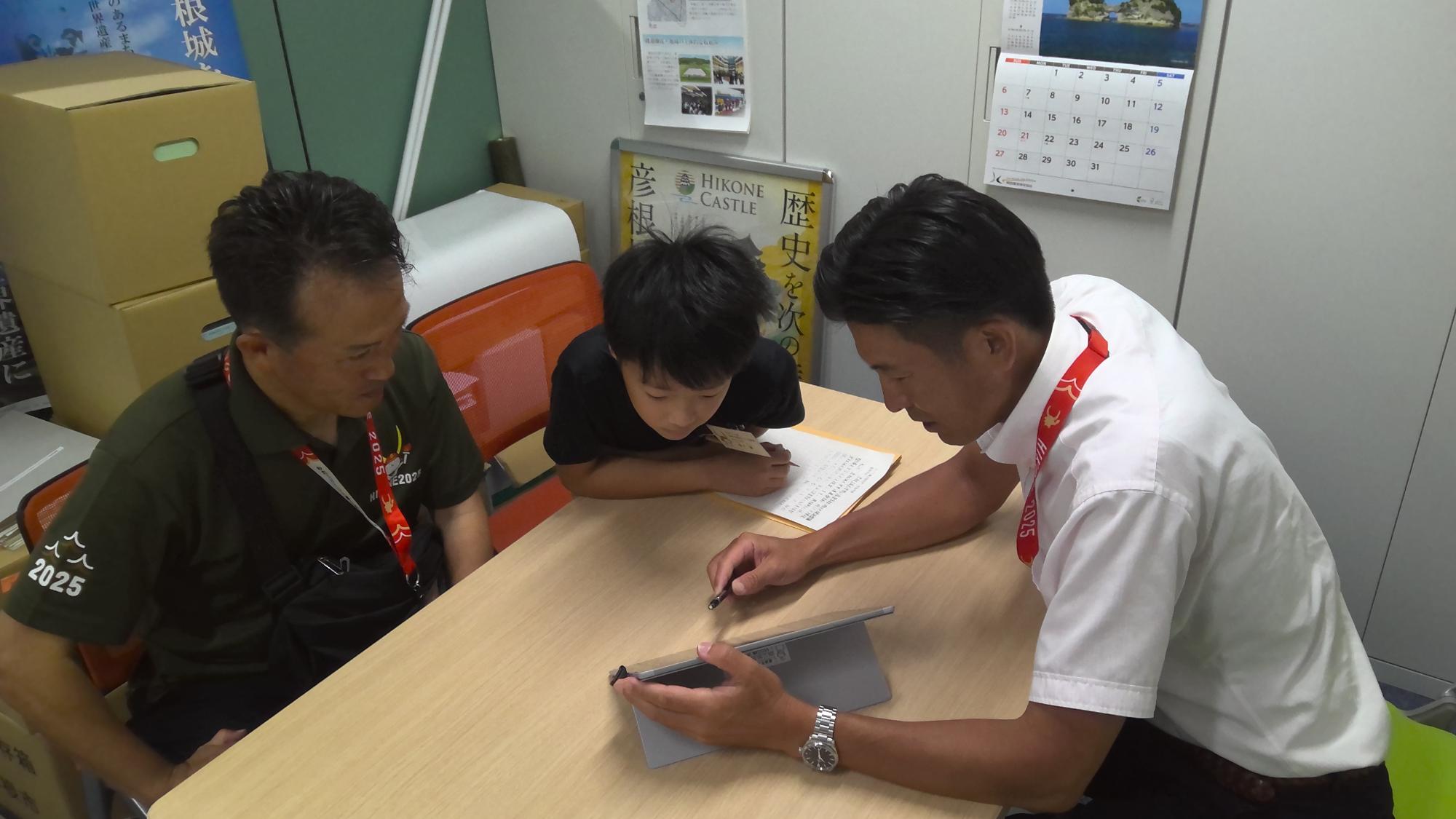 タブレットを使用し説明する職員と、のぞき込む子供議員の写真