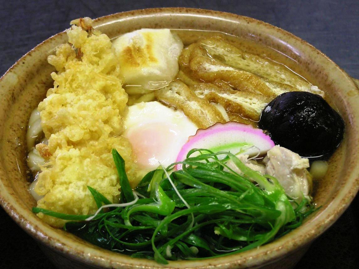 油揚げ、天ぷら、シイタケ、半熟卵、餅などが入った(しる万本店)のうどんの写真
