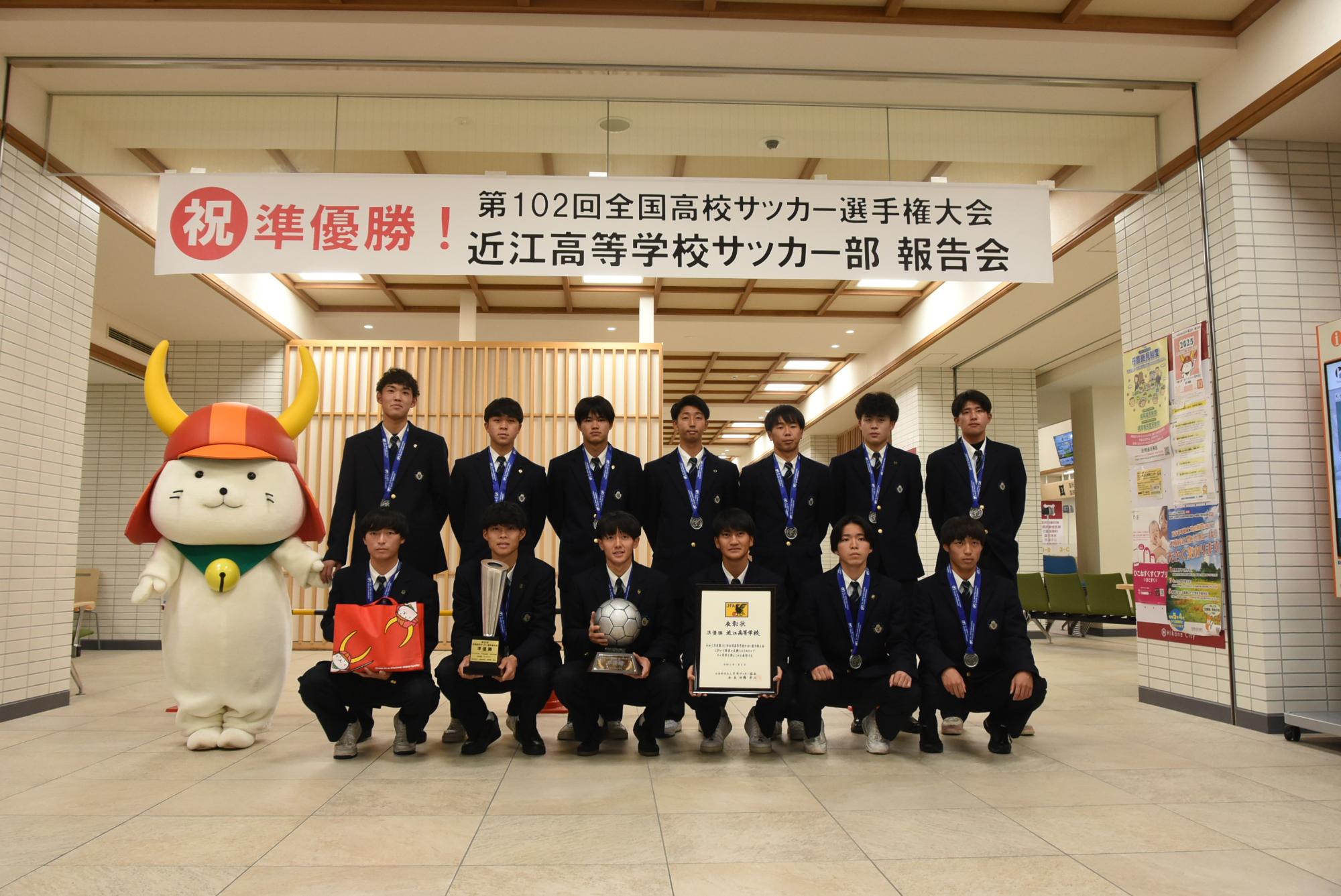 横断幕の下でひこにゃんとトロフィー等を持った選手達の集合写真