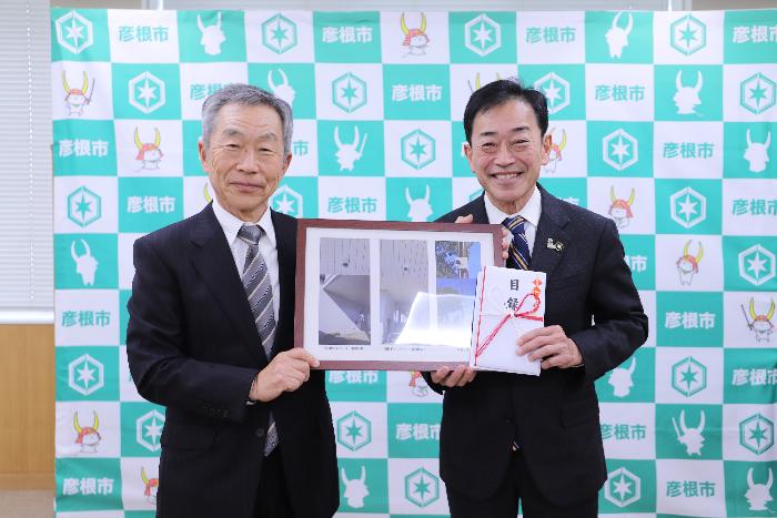 防犯カメラ寄付受領式で目録や写真を持ち記念撮影する市長と滋賀県遊技業行動組合彦根支部の男性