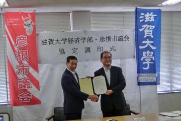 滋賀大学経済学部と彦根市議会の方が調印書を持ち握手をしている写真