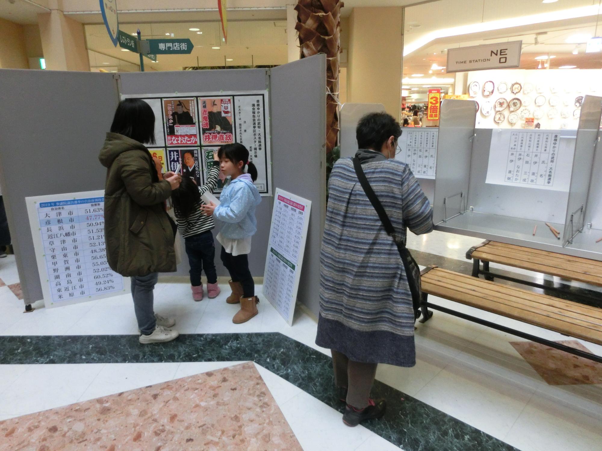 模擬選挙の選挙ポスターを見る親子と投票用紙に記入をしている女性の写真