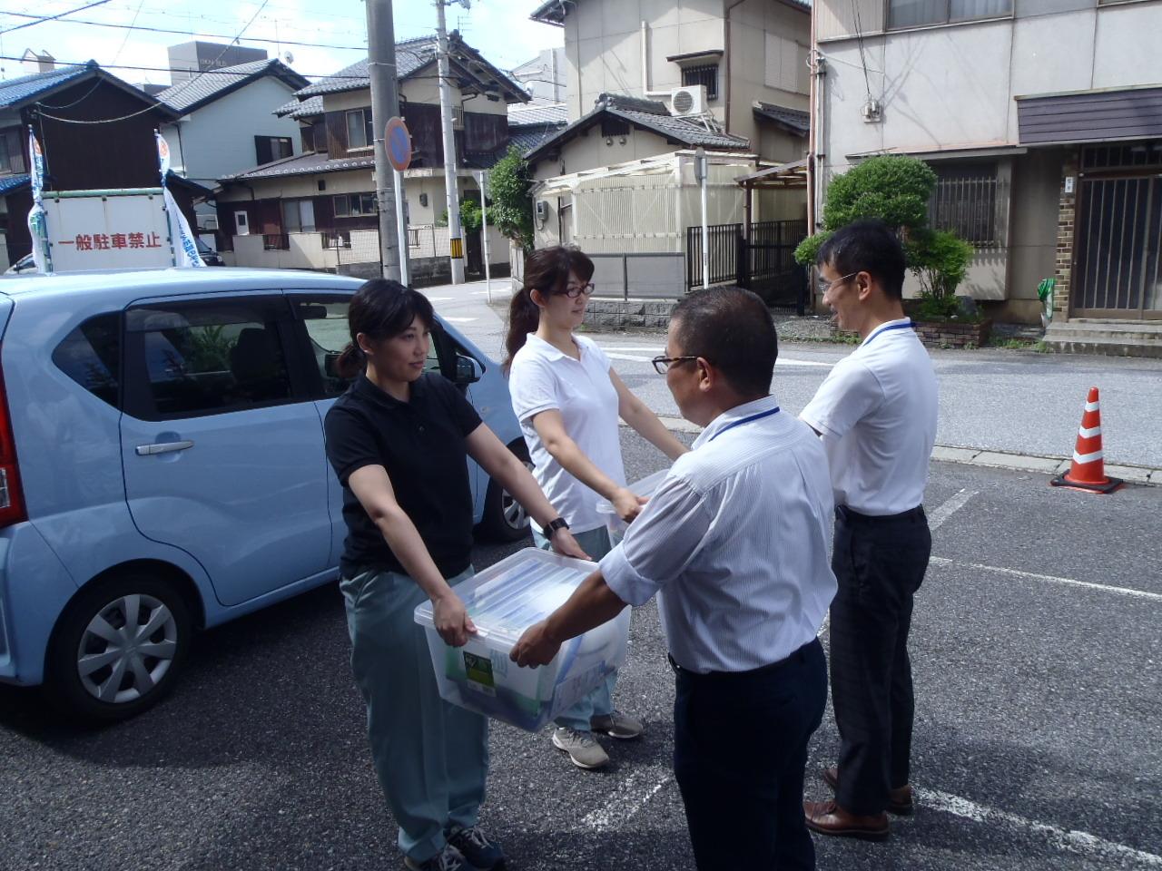 駐車場にて車を後ろに男性2名から荷物を受け取る女性2名の写真