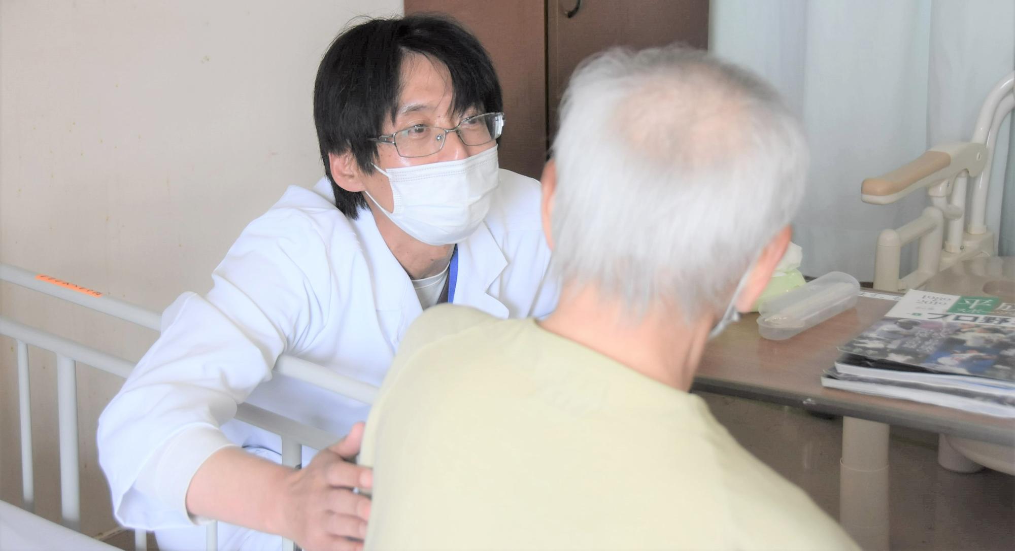 病室で、かがみこんで患者さんの背中にそっと手を添える白衣の医師