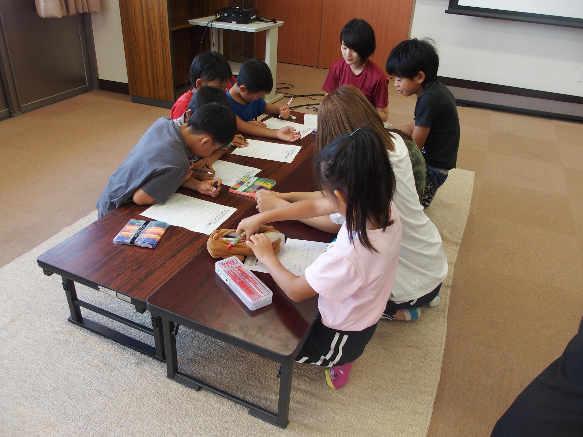 机を前に正座した7人の子供に女性指導員二人が勉強を教えている写真