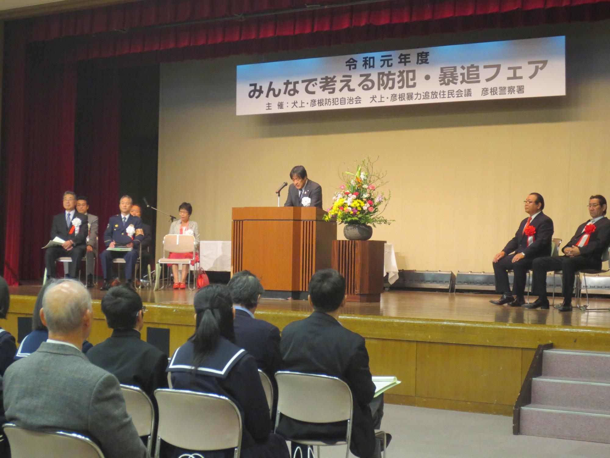 会場の様子（壇上で話す話者と聴衆、来賓）の写真