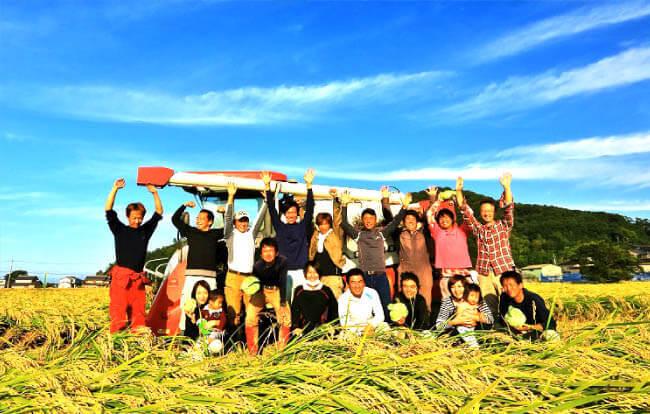 晴れた秋空の下収穫を迎えた稲の田んぼの中で万歳をする農家とその家族の写真