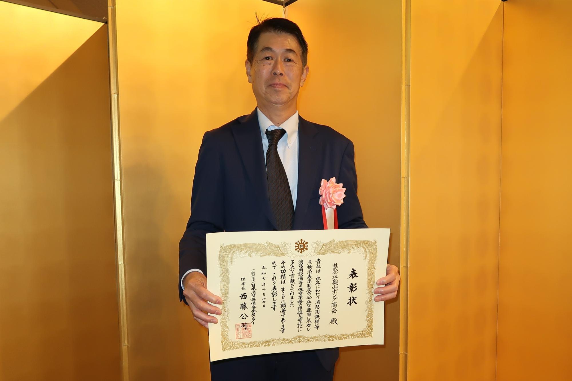 男性が金屏風の前で表彰状をもっている写真