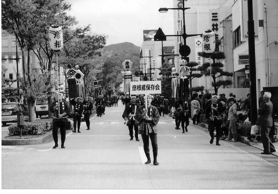 彦根鳶保存会のプラカードを持ち法被を着て街頭を歩いている保存会の方々の白黒写真