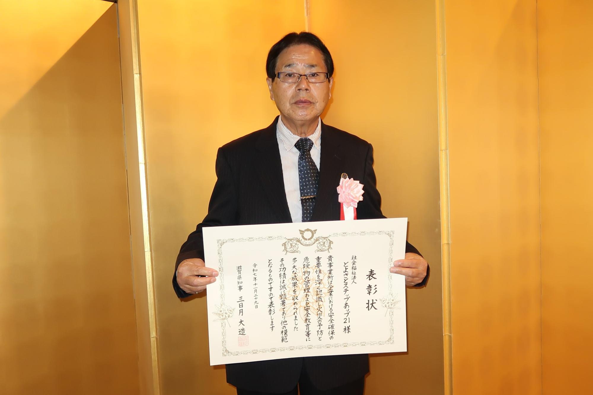 男性が金屏風の前で表彰状をもっている写真