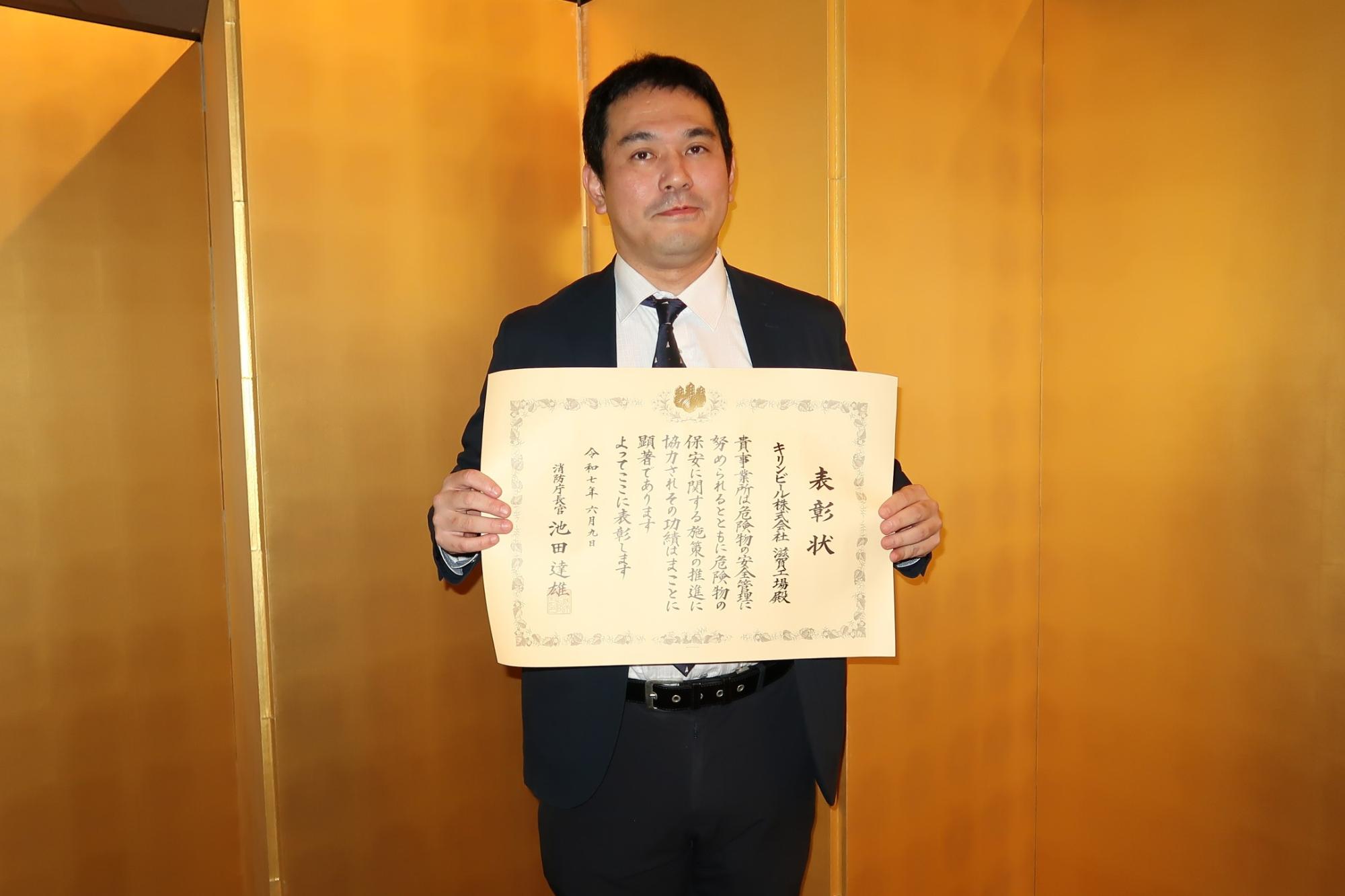 男性が金屏風の前で表彰状をもっている写真